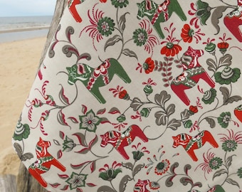 May include: Cream-colored fabric with a repeating pattern of red and green Dala horses and floral designs. The fabric is draped over a wooden post, with a beach and ocean in the background.