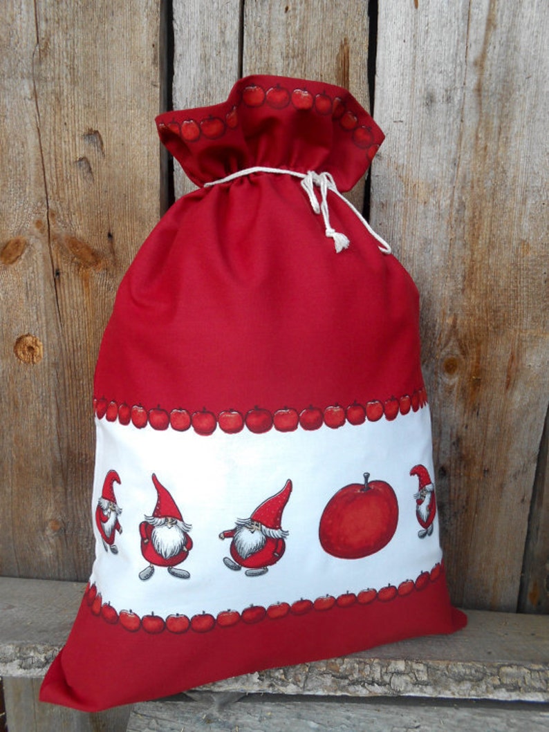 May include: Red fabric gift bag with a white border featuring a row of red apples and gnomes. The bag has a drawstring closure.