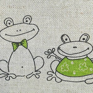Puede incluir: Dos ranas de dibujos animados sobre un fondo de tela beige. La rana m&aacute;s grande lleva una corbata de lazo verde y la rana m&aacute;s peque&ntilde;a lleva una camisa verde con un patr&oacute;n de tr&eacute;boles verdes.