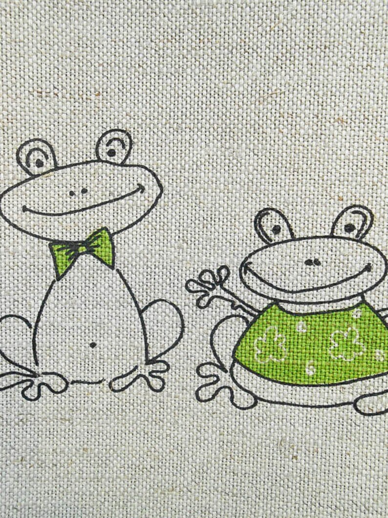 Puede incluir: Dos ranas de dibujos animados sobre un fondo de tela beige. La rana m&aacute;s grande lleva una corbata de lazo verde y la rana m&aacute;s peque&ntilde;a lleva una camisa verde con un patr&oacute;n de tr&eacute;boles verdes.