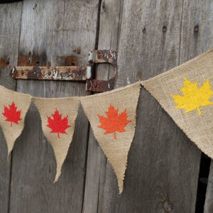 May include: A burlap banner with a string of triangular flags. Each flag features a different colored maple leaf, including red, orange, and yellow.