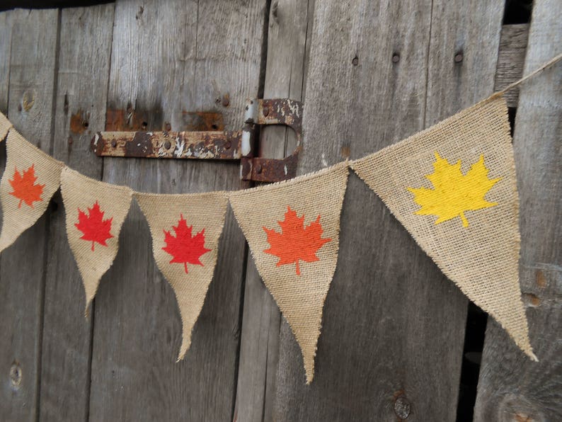 May include: A burlap banner with a string of triangular flags. Each flag features a different colored maple leaf, including red, orange, and yellow.
