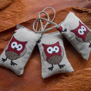 May include: Three small, gray fabric owl-shaped sachets with red and brown accents. The owls have black eyes and beaks. The sachets are tied together with a white string.
