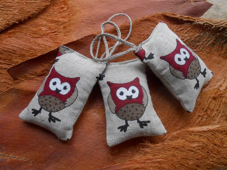 May include: Three small, gray fabric owl-shaped sachets with red and brown accents. The owls have black eyes and beaks. The sachets are tied together with a white string.