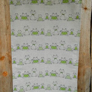 Puede incluir: Un pa&ntilde;o de cocina de lino con un patr&oacute;n repetido de ranas verdes con contornos negros. Las ranas est&aacute;n en varias poses, algunas con pajaritas, otras con las piernas extendidas y otras con la boca abierta. El pa&ntilde;o est&aacute; colgado en una superficie de madera.