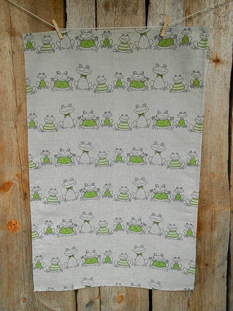 Puede incluir: Un pa&ntilde;o de cocina de lino con un patr&oacute;n repetido de ranas verdes con contornos negros. Las ranas est&aacute;n en varias poses, algunas con pajaritas, otras con las piernas extendidas y otras con la boca abierta. El pa&ntilde;o est&aacute; colgado en una superficie de madera.