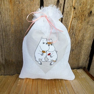 May include: White fabric gift bag with a pink ribbon and a heart-shaped design featuring two cartoon characters. The characters are white with black outlines and are holding a flower.