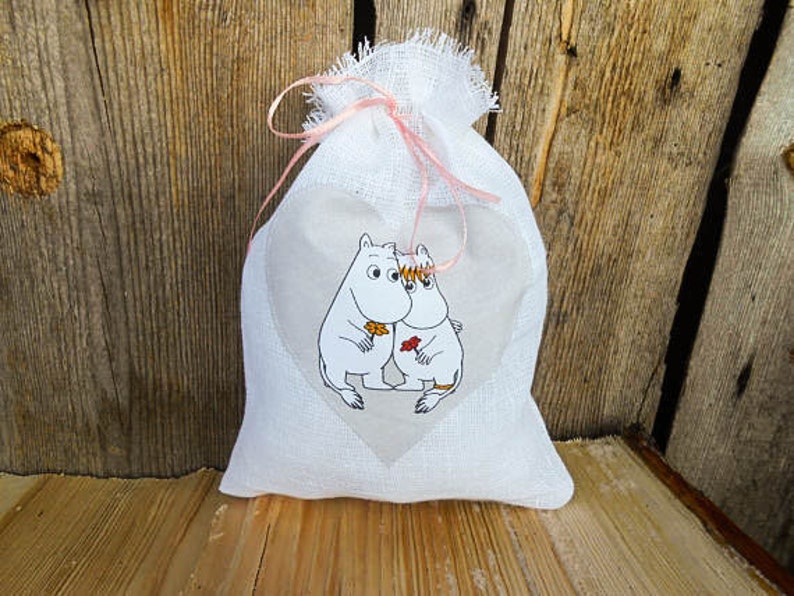 May include: White fabric gift bag with a pink ribbon and a heart-shaped design featuring two cartoon characters. The characters are white with black outlines and are holding a flower.