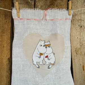 May include: A white fabric bag with a pink ribbon and a heart-shaped patch featuring two cartoon characters. The characters are white with orange and brown details and are holding flowers.