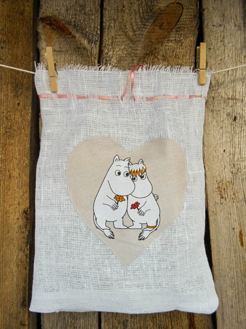 May include: A white fabric bag with a pink ribbon and a heart-shaped patch featuring two cartoon characters. The characters are white with orange and brown details and are holding flowers.