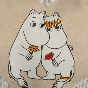 May include: Two white cartoon characters with black outlines and orange and red flowers are hugging on a beige fabric background. The characters have large black eyes and are holding flowers.