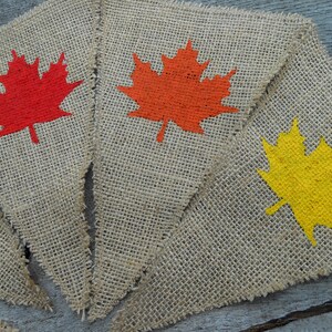 May include: Three burlap pennants with painted maple leaves. The leaves are red, orange, and yellow.