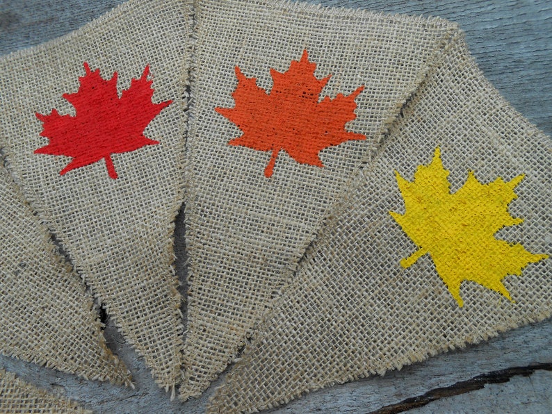 May include: Three burlap pennants with painted maple leaves. The leaves are red, orange, and yellow.