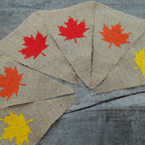 May include: A burlap banner with six triangular flags, each featuring a different colored maple leaf. The leaves are painted in shades of red, orange, and yellow.