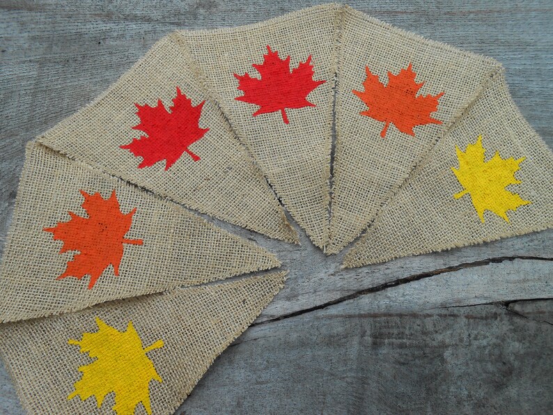 May include: A burlap banner with six triangular flags, each featuring a different colored maple leaf. The leaves are painted in shades of red, orange, and yellow.