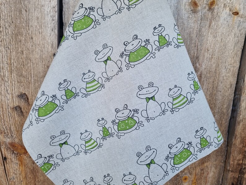 Puede incluir: Un pa&ntilde;o de cocina de lino blanco con un patr&oacute;n de ranas verdes. Las ranas llevan diferentes atuendos, incluyendo una corbata de lazo, una camisa a rayas y un su&eacute;ter.