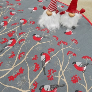 May include: A gray and red Christmas tree skirt with a white crocheted trim. The skirt features a repeating pattern of red berries, branches, and birds. Two gray gnomes with red hats are placed on the skirt.