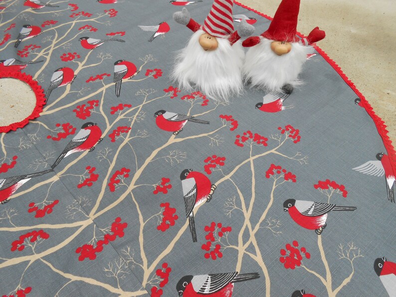 May include: A gray and red Christmas tree skirt with a white crocheted trim. The skirt features a repeating pattern of red berries, branches, and birds. Two gray gnomes with red hats are placed on the skirt.