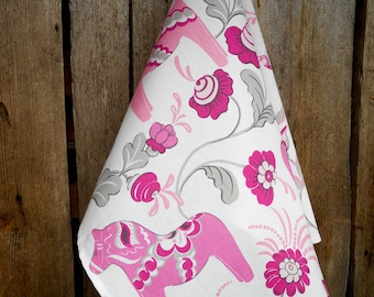 May include: A white tea towel with a pink and gray floral pattern featuring stylized horses. The horses are pink with gray and white details. The flowers are pink with white centers.