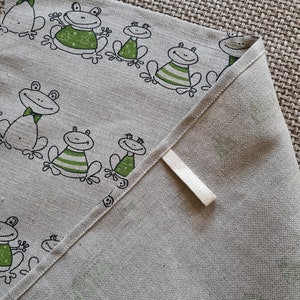 Puede incluir: Un pa&ntilde;o de cocina de lino beige con un dise&ntilde;o de rana verde y negra. La toalla tiene un lazo de colgar blanco.