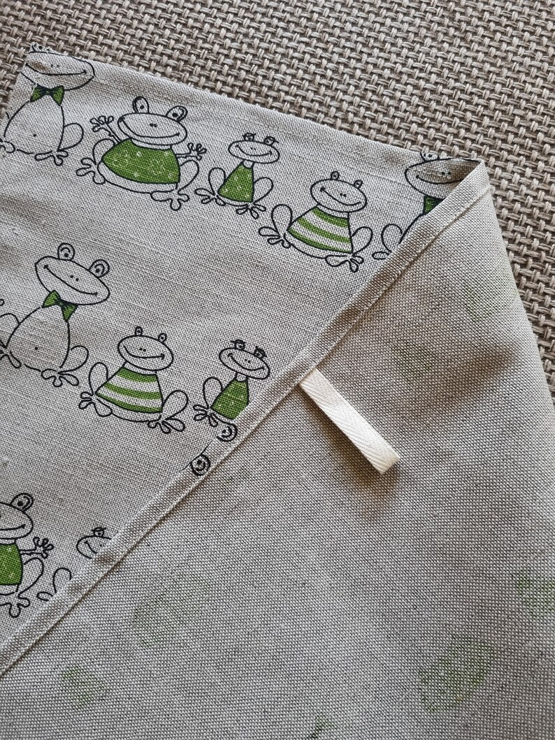 Puede incluir: Un pa&ntilde;o de cocina de lino beige con un dise&ntilde;o de rana verde y negra. La toalla tiene un lazo de colgar blanco.