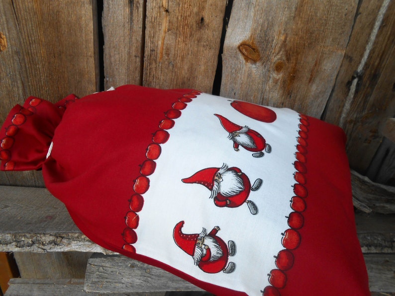 May include: Red and white fabric bag with a drawstring closure. The bag is decorated with a pattern of red apples and gnomes.