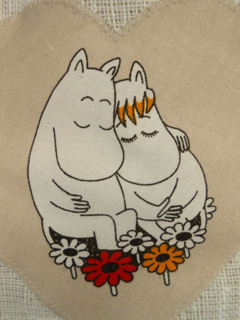 May include: Two white cartoon characters with black outlines are hugging each other. They are surrounded by five colorful flowers with black centers. The background is a beige fabric with a white lace border.