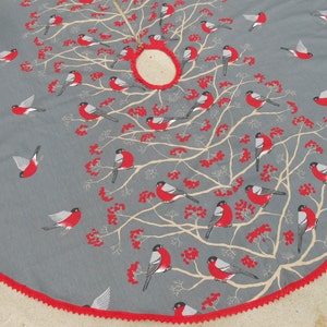 May include: A gray and red Christmas tree skirt with a white tree design and red birds. The skirt has a red scalloped edge.