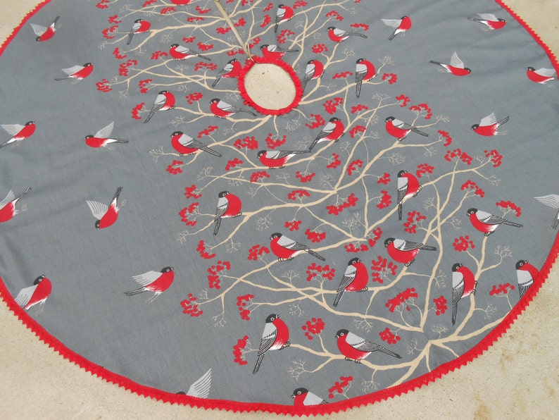 May include: A gray and red Christmas tree skirt with a white tree design and red birds. The skirt has a red scalloped edge.