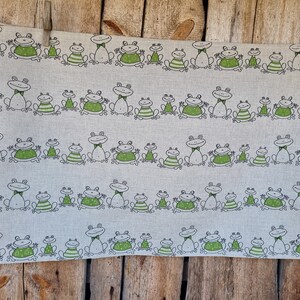 Puede incluir: Una toalla de t&eacute; de lino blanco con un patr&oacute;n repetido de ranas verdes. Las ranas est&aacute;n dibujadas en un estilo simple y de dibujos animados y llevan pajaritas. La toalla est&aacute; colgada en una valla de madera.