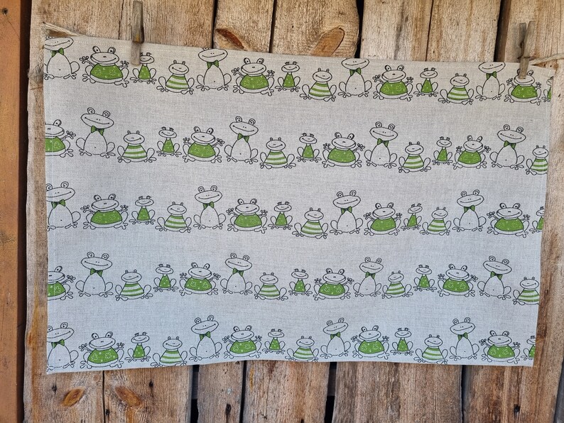 Puede incluir: Una toalla de t&eacute; de lino blanco con un patr&oacute;n repetido de ranas verdes. Las ranas est&aacute;n dibujadas en un estilo simple y de dibujos animados y llevan pajaritas. La toalla est&aacute; colgada en una valla de madera.
