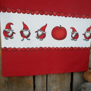 May include: Red and white kitchen towel with a border of red apples and a row of gnomes in red hats. The gnomes are holding apples and walking in a line.