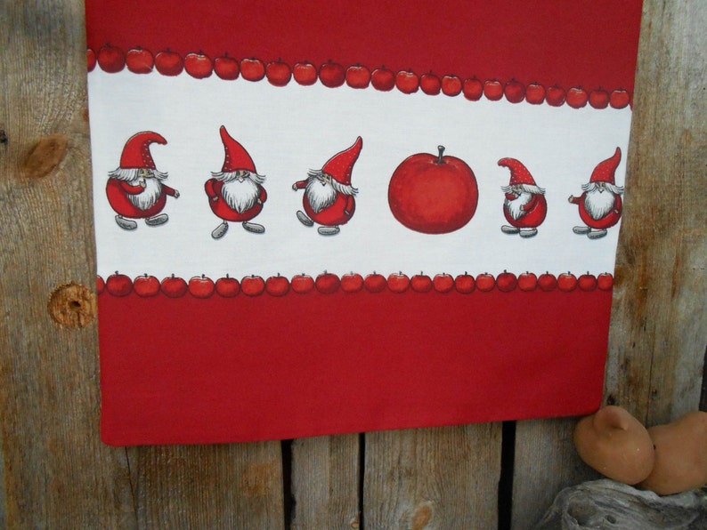 May include: Red and white kitchen towel with a border of red apples and a row of gnomes in red hats. The gnomes are holding apples and walking in a line.