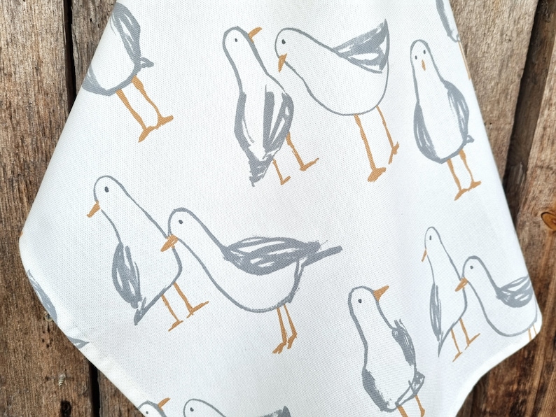 Puede incluir: Tela blanca con un patr&oacute;n repetido de gaviotas grises estilizadas con picos y patas naranjas. Las aves est&aacute;n esbozadas en un estilo simple e ilustrativo, creando un dise&ntilde;o caprichoso. La tela es probablemente un pa&ntilde;o de cocina o similar.