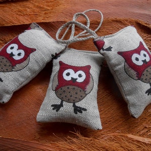 May include: Three gray linen owl-shaped sachets with red and brown accents. The owls have large eyes and are holding their wings out.