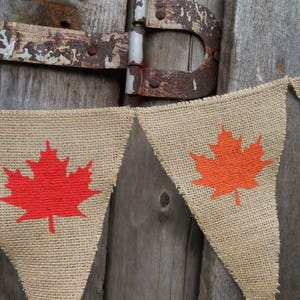 May include: A burlap banner with orange and red maple leaf designs. The banner is strung on a rustic wooden background.