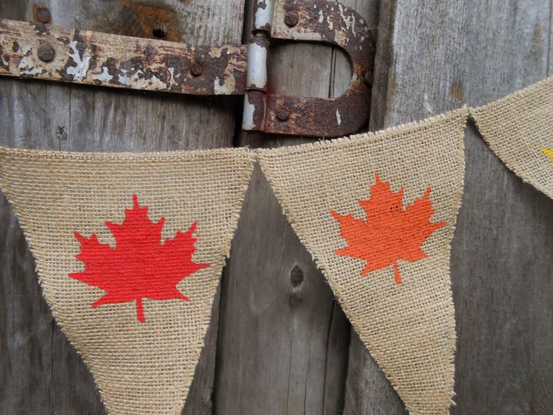 May include: A burlap banner with orange and red maple leaf designs. The banner is strung on a rustic wooden background.