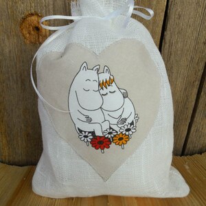 May include: A white linen drawstring bag with a heart-shaped patch featuring a cartoon illustration of two characters hugging. The characters are white with black outlines and have closed eyes. The heart-shaped patch is surrounded by colorful flowers.
