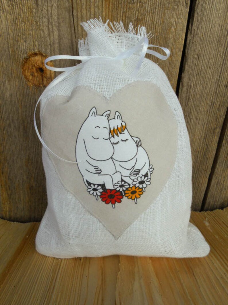May include: A white linen drawstring bag with a heart-shaped patch featuring a cartoon illustration of two characters hugging. The characters are white with black outlines and have closed eyes. The heart-shaped patch is surrounded by colorful flowers.