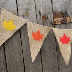 May include: A string of burlap pennants with a yellow maple leaf, a red maple leaf, and two more red maple leaves. The pennants are hanging on a wooden wall.