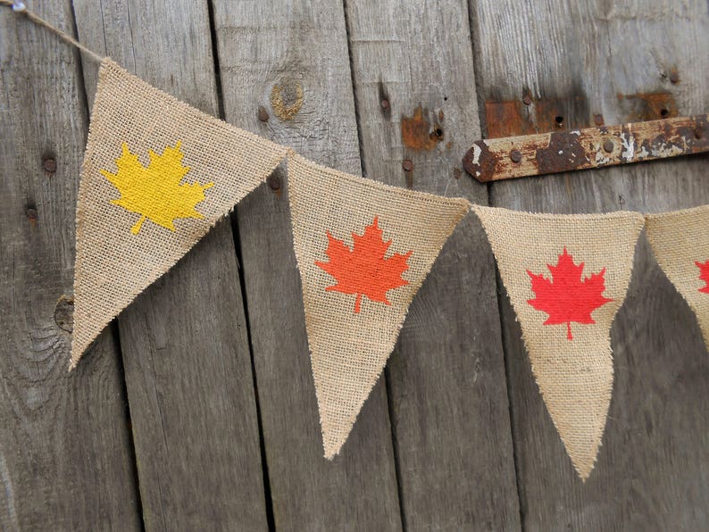 May include: A string of burlap pennants with a yellow maple leaf, a red maple leaf, and two more red maple leaves. The pennants are hanging on a wooden wall.