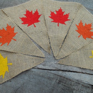 May include: A burlap banner with six triangular flags, each featuring a different colored maple leaf. The leaves are red, orange, and yellow.