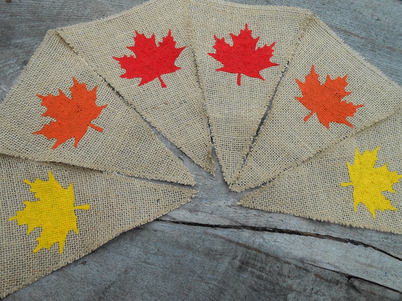 May include: A burlap banner with six triangular flags, each featuring a different colored maple leaf. The leaves are red, orange, and yellow.