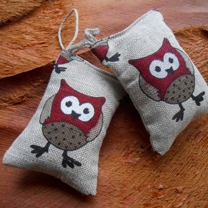 May include: Two small, beige fabric pouches with red and black owl designs. The owls have large, round eyes and are perched on a brown surface.