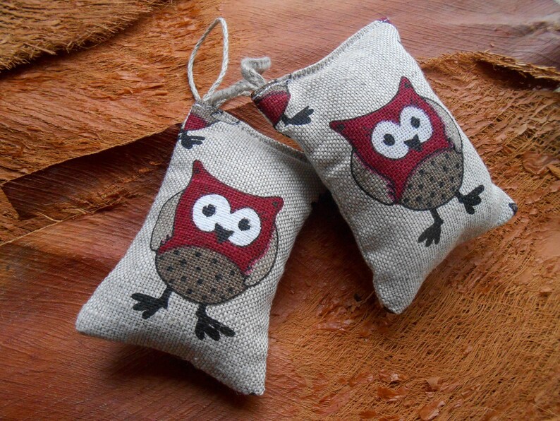 May include: Two small, beige fabric pouches with red and black owl designs. The owls have large, round eyes and are perched on a brown surface.