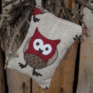 May include: A small, beige fabric pouch with a red owl design. The owl has black eyes and a white beak. The pouch is hanging from a brown string.