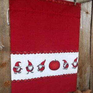 May include: Red fabric drawstring bag with a white border featuring a row of red apples and a row of gnomes with red hats. The bag is hanging from a wooden surface with clothespins.