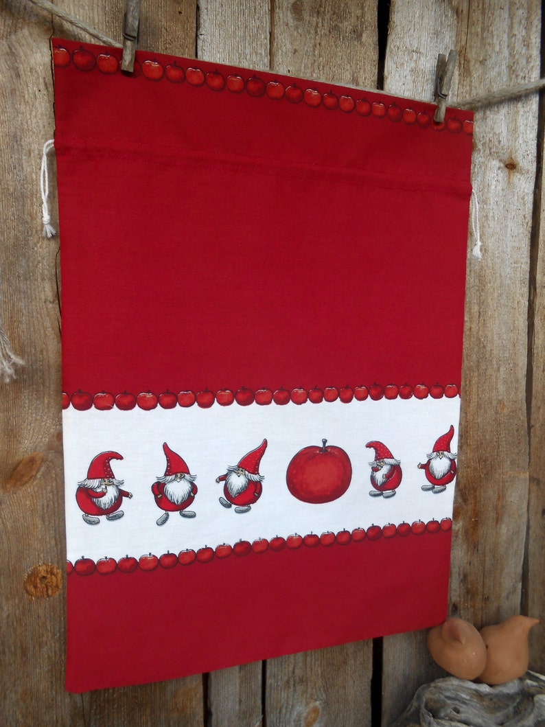 May include: Red fabric drawstring bag with a white border featuring a row of red apples and a row of gnomes with red hats. The bag is hanging from a wooden surface with clothespins.