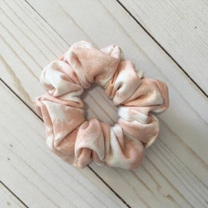 May include: A scrunchie with a tie-dye pattern in shades of peach and white. The fabric is gathered and elasticized, creating a ruffled appearance. This hair accessory is designed to hold hair in a ponytail or bun.