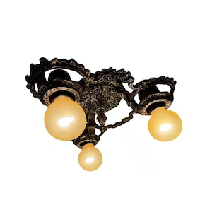 May include: Ornate bronze-colored ceiling light fixture with three spherical light bulbs. The fixture has a detailed, textured design with swirling patterns and a dark finish. The bulbs emit a warm, yellow glow, creating a vintage aesthetic.